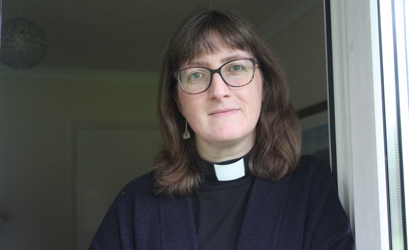 Kate Massey (a woman in a clerical collar with brown shoulder length hair and glasses) standing at a doorway looking at the camera. doorway