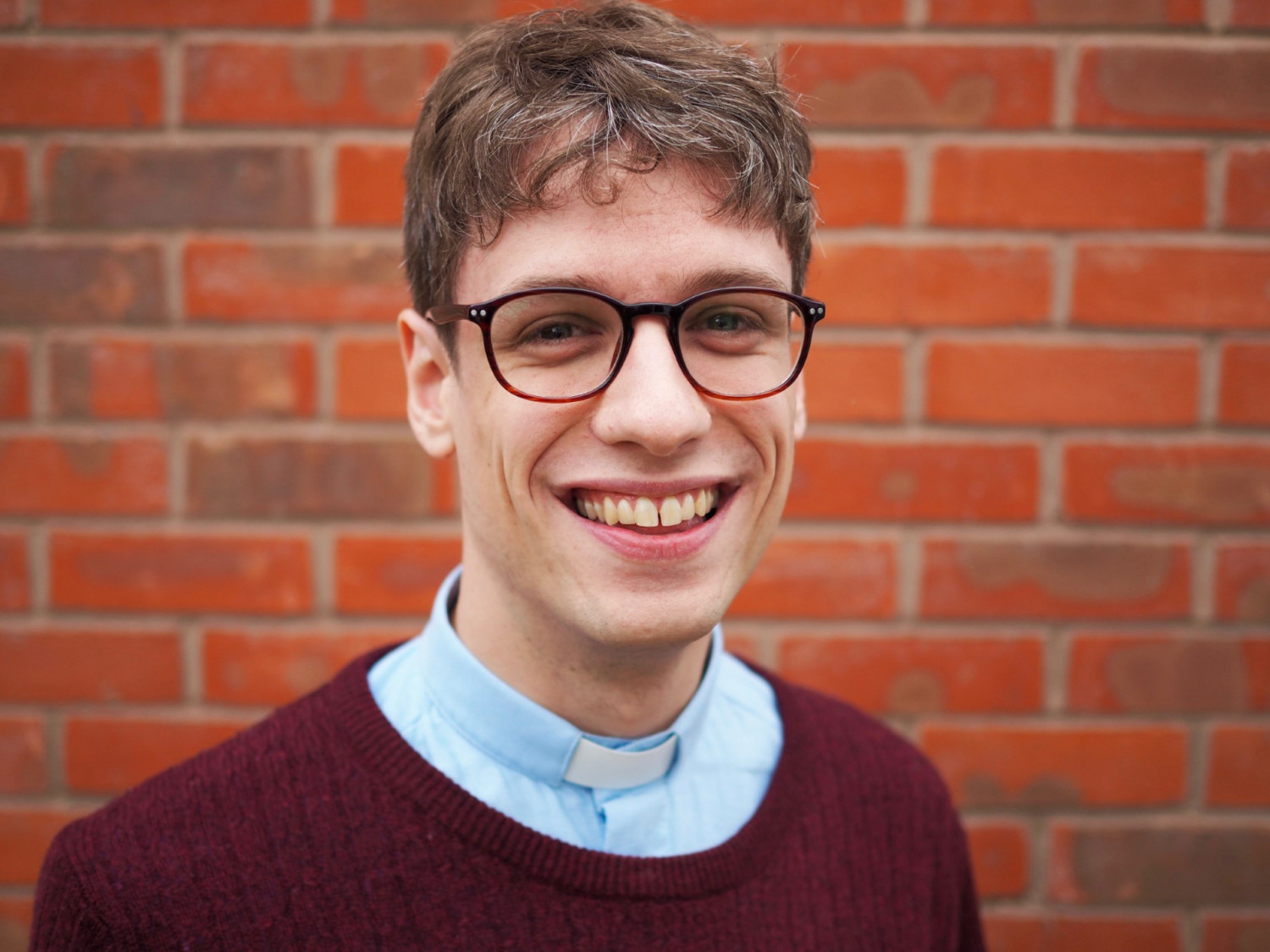 Charles, a white man wearing glasses with dark blonde hair standing in front of a brick wall and smiling